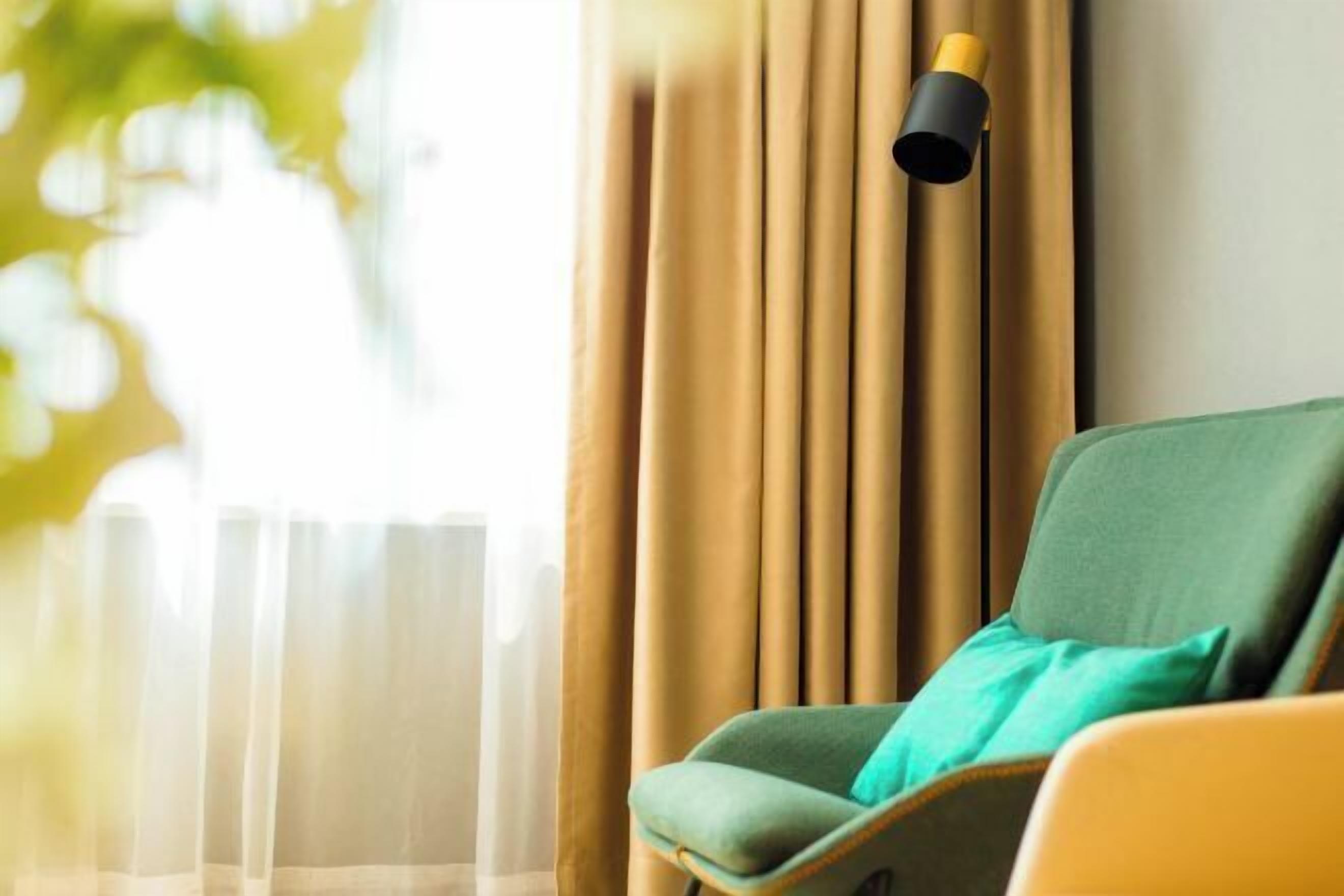 A room with a green armchair, a black and yellow lamp, and beige curtains.
