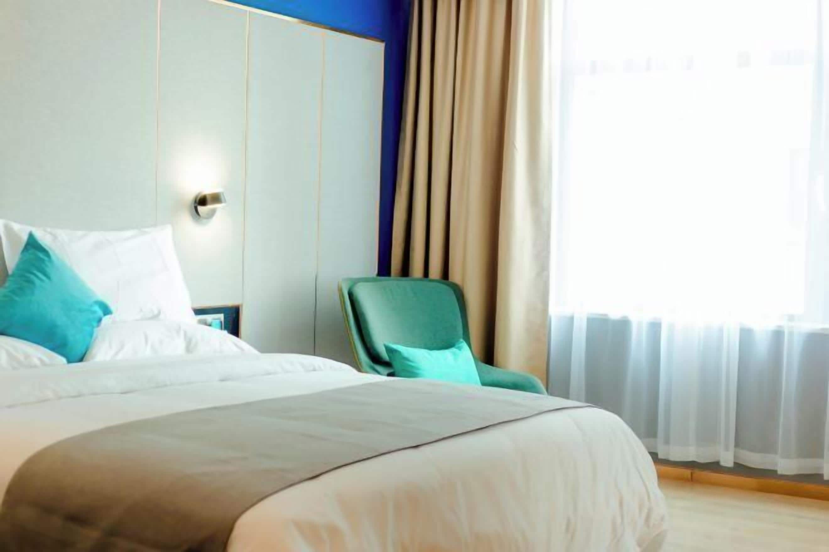 Ett hotellrum med en stor säng, en grön fåtölj och utsikt över omgivningarna genom tunna gardiner.