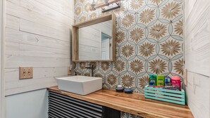 Ein Badezimmer mit einem Holzwaschtisch, einem weißen Waschbecken, einem Spiegel und einer dekorativen Fliesenrückwand.