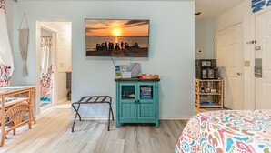 Ein Zimmer mit einem türkisfarbenen Schrank, einem Fernseher, der eine Sonnenuntergangsszene zeigt, einem Korbstuhl und einem Bett mit einer bunten Decke.
