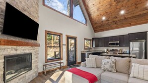 A modern living room with a fireplace, a sofa, and a kitchen area with dark cabinets and stainless steel appliances. - Anchor Point- Hot Tub, Firepit, Ping Pong (Rogers)
