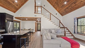 A spacious kitchen with a large island, a dining area, a living room with a sofa, and a wooden staircase. - Anchor Point- Hot Tub, Firepit, Ping Pong (Rogers)
