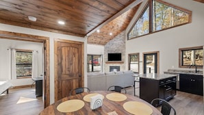 A spacious kitchen with a stone fireplace, a dining area with a wooden table, and large windows offering views of the outdoors. - Anchor Point- Hot Tub, Firepit, Ping Pong (Rogers)