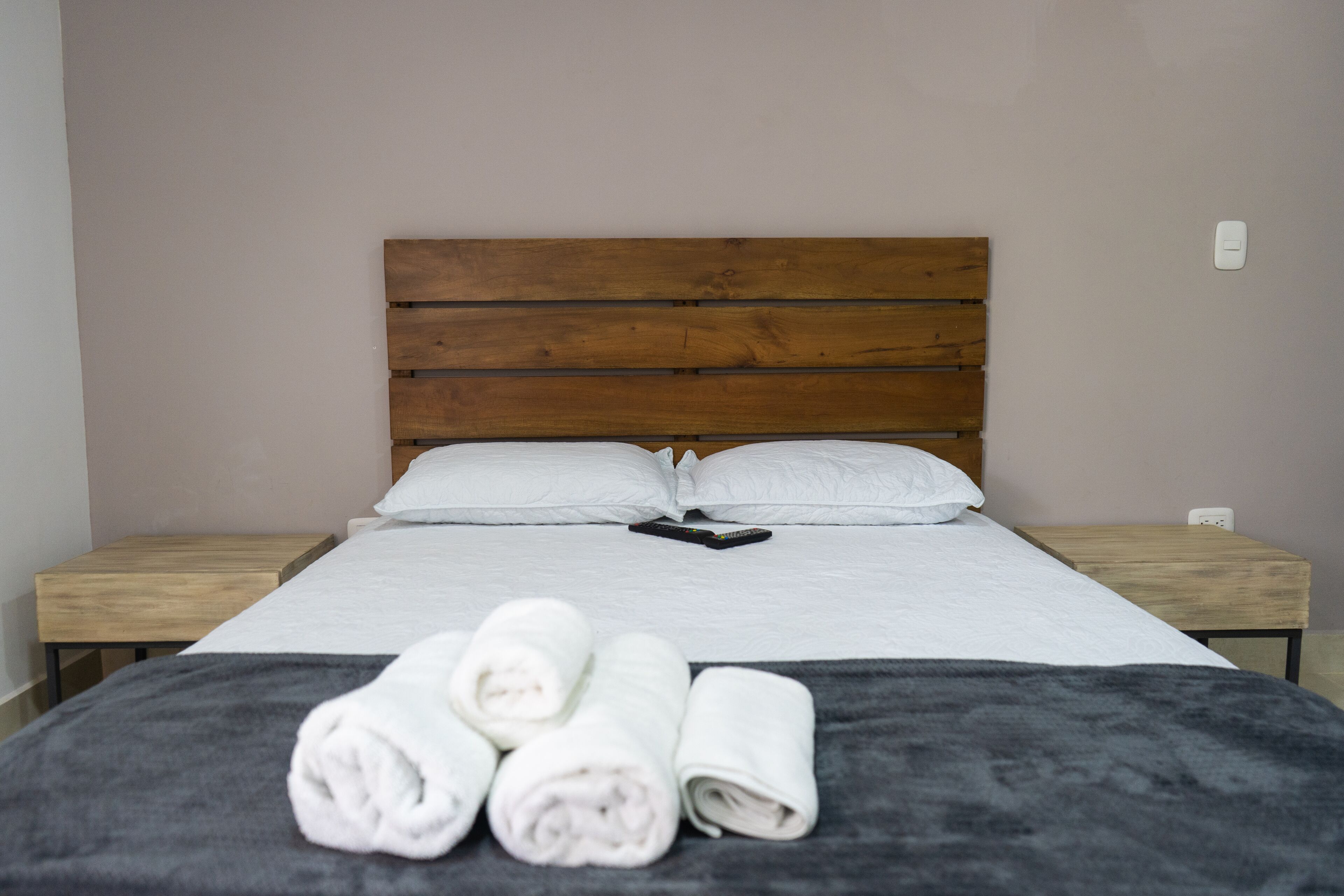 A neatly made bed with white linens, a wooden headboard, and a remote control on the bed. There are rolled towels on the bed and a small wooden nightstand beside the bed.