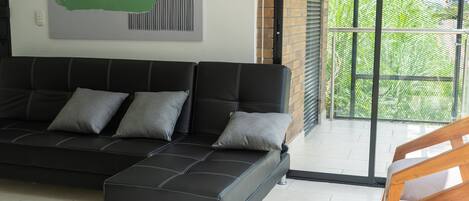 Un salon moderne avec un canapé d’angle noir, un fauteuil en bois et une grande fenêtre donnant sur un paysage verdoyant.