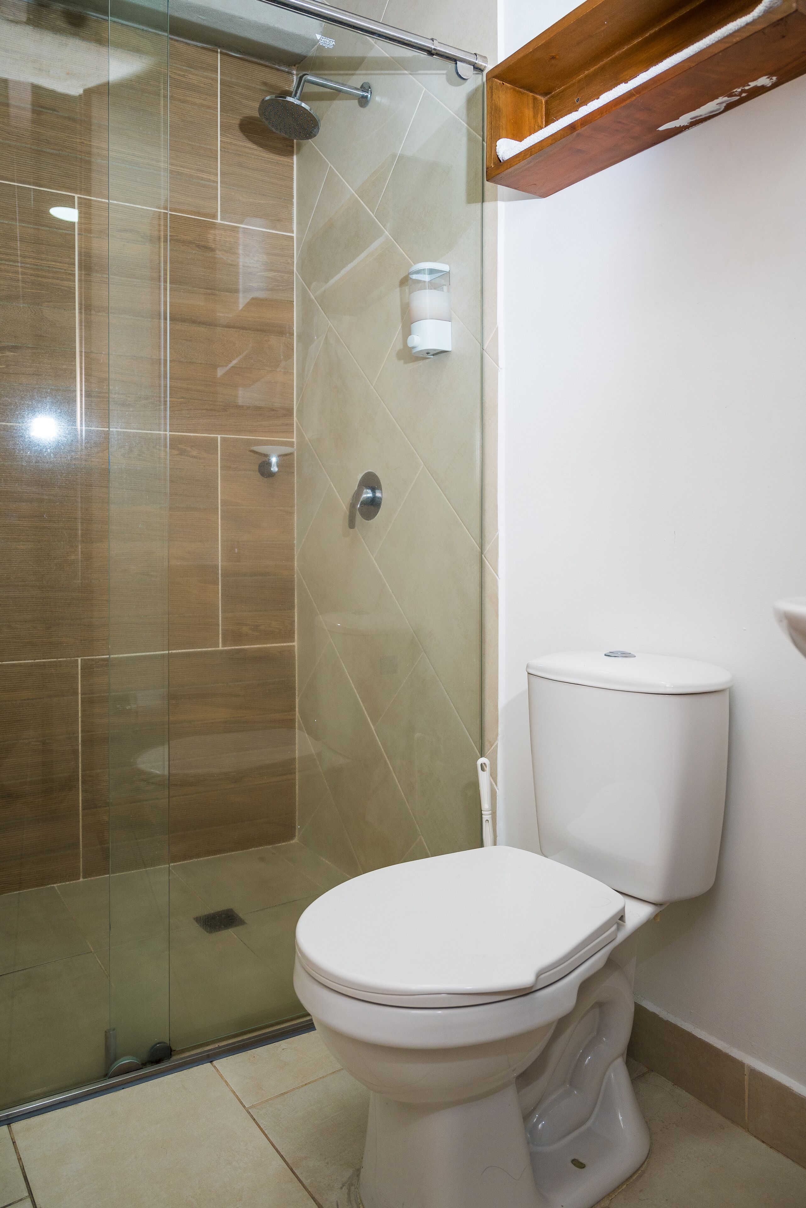 A bathroom with a glass-enclosed shower, a white toilet, and a wooden shelf above the toilet.