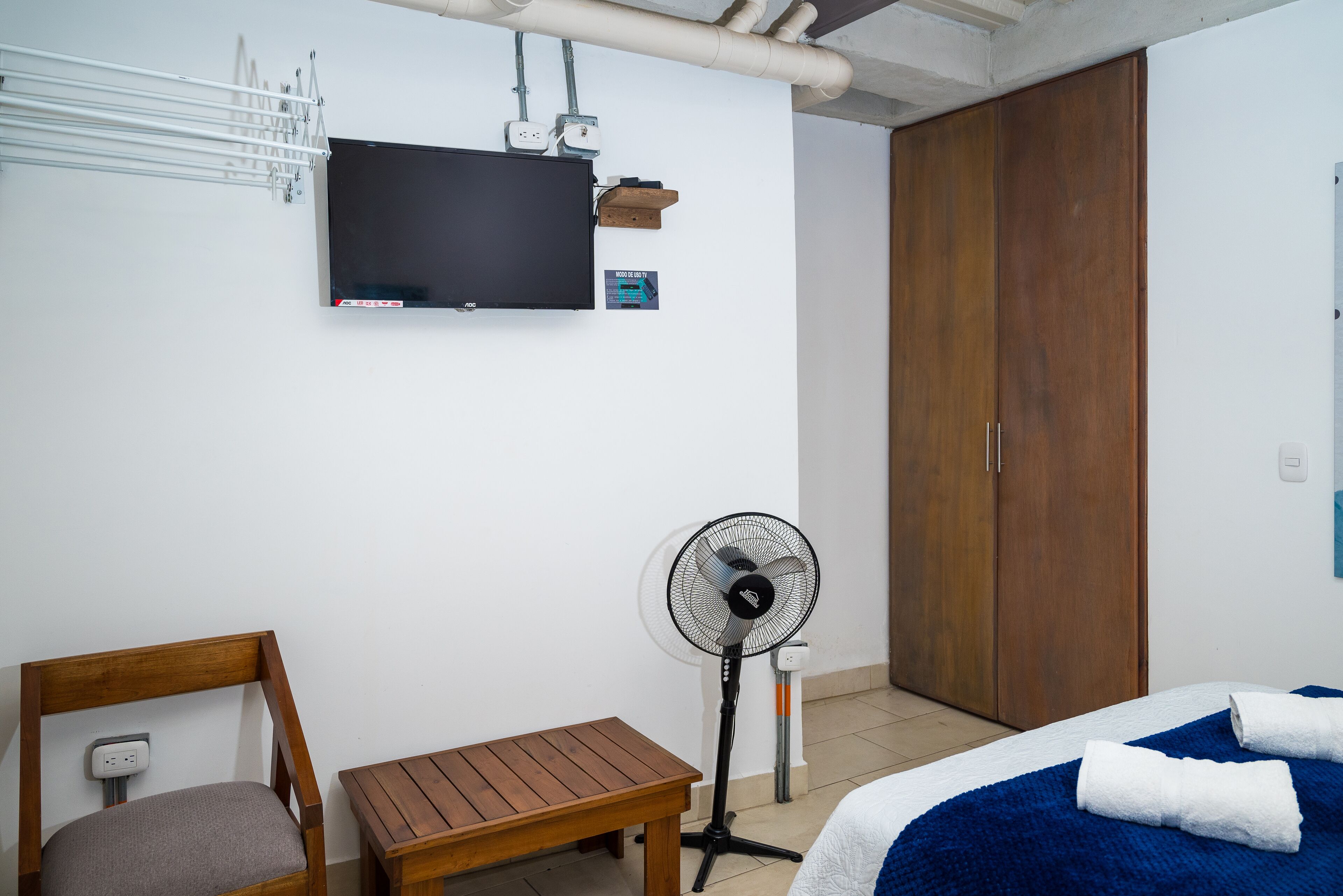 Una habitación de hotel con una cama, una silla de madera, una mesita, un ventilador de pie, un televisor montado en la pared y un armario.