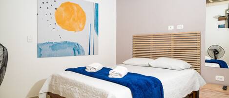 Una camera da letto con un letto, una coperta blu, cuscini bianchi, una testiera in legno, un quadro al muro, una ventola e un piccolo tavolo.