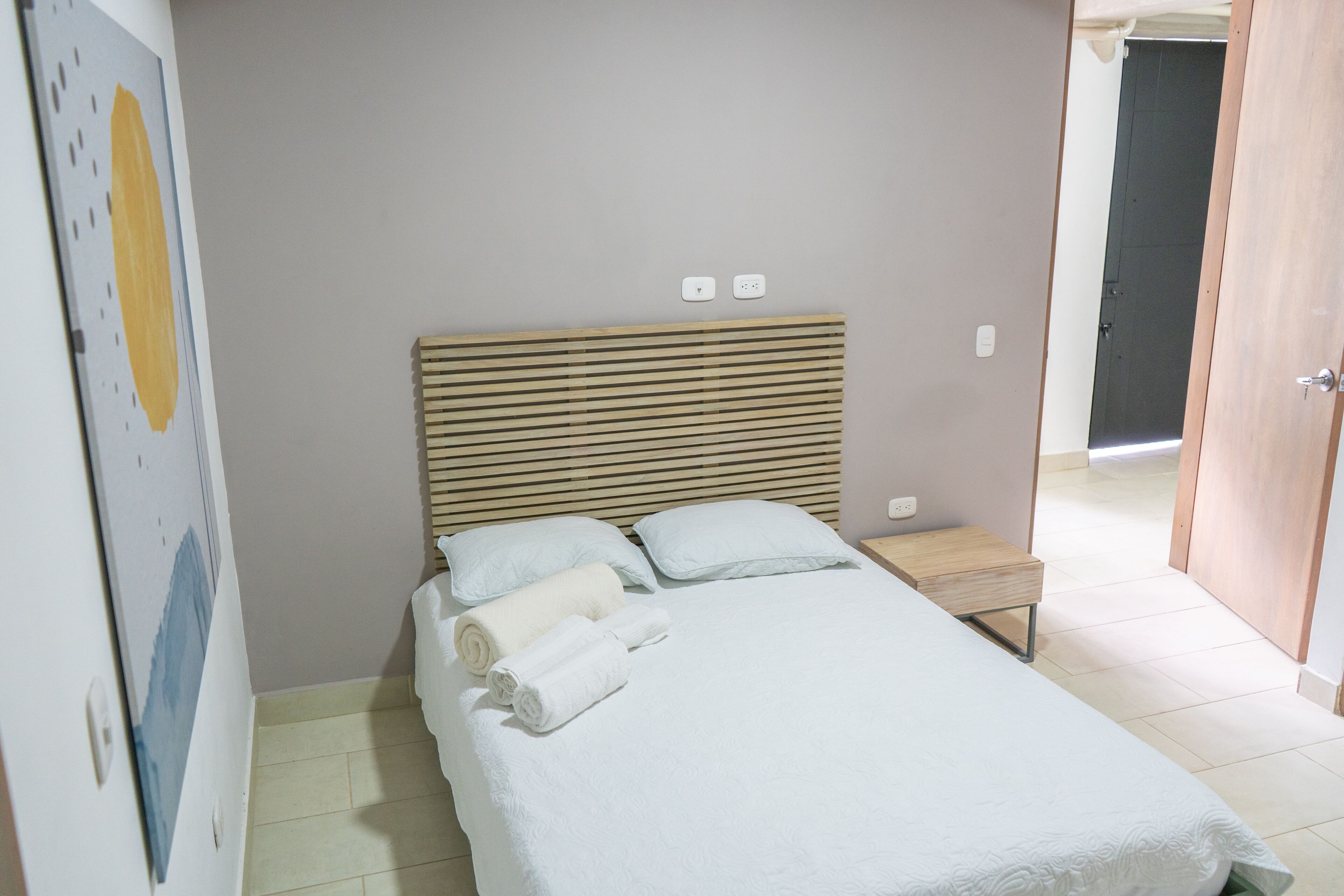A neatly made bed with pillows and rolled towels, a bedside table, and a wall-mounted light switch.