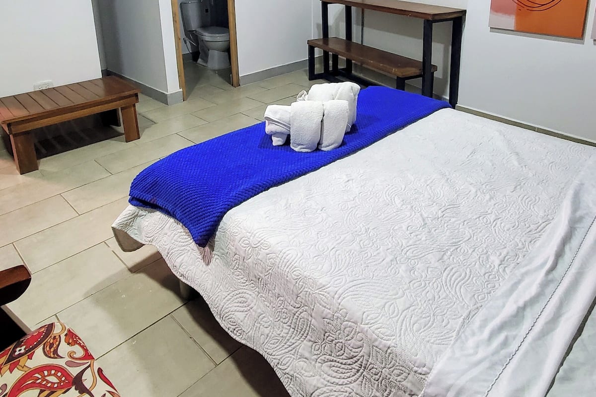 Quarto com cama, um banco de madeira, uma mesa pequena e um banheiro visível por uma porta aberta.