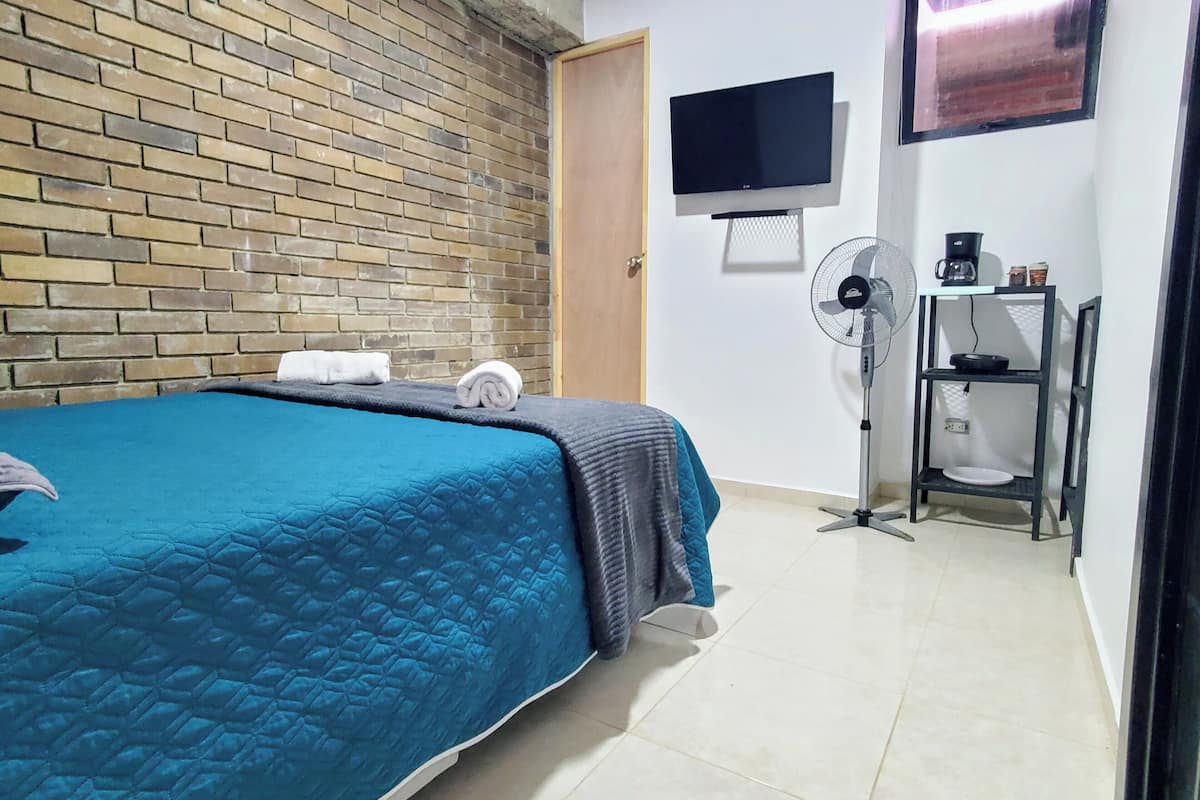Quarto de hotel com parede em padrão tijolo, cama com colcha azul, televisão de tela plana, ventilador e uma pequena mesa com cafeteira e um prato.