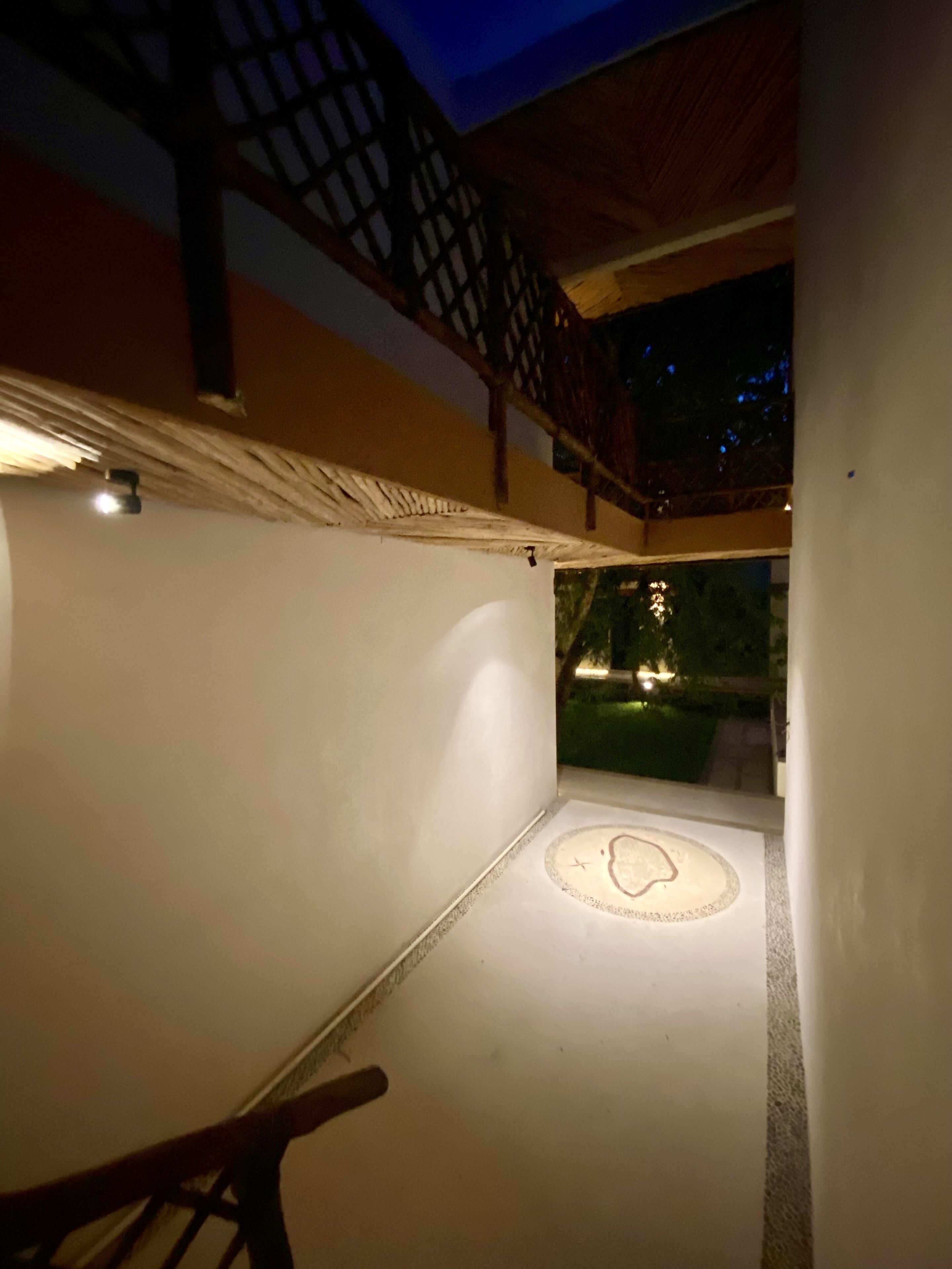 Un espacio interior tenuemente iluminado, con suelo estampado, una escalera y vista a un jardín exterior de noche.