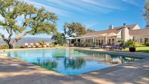 Una piscina con sillas de descanso y sombrillas, rodeada de árboles y una casa con un patio.