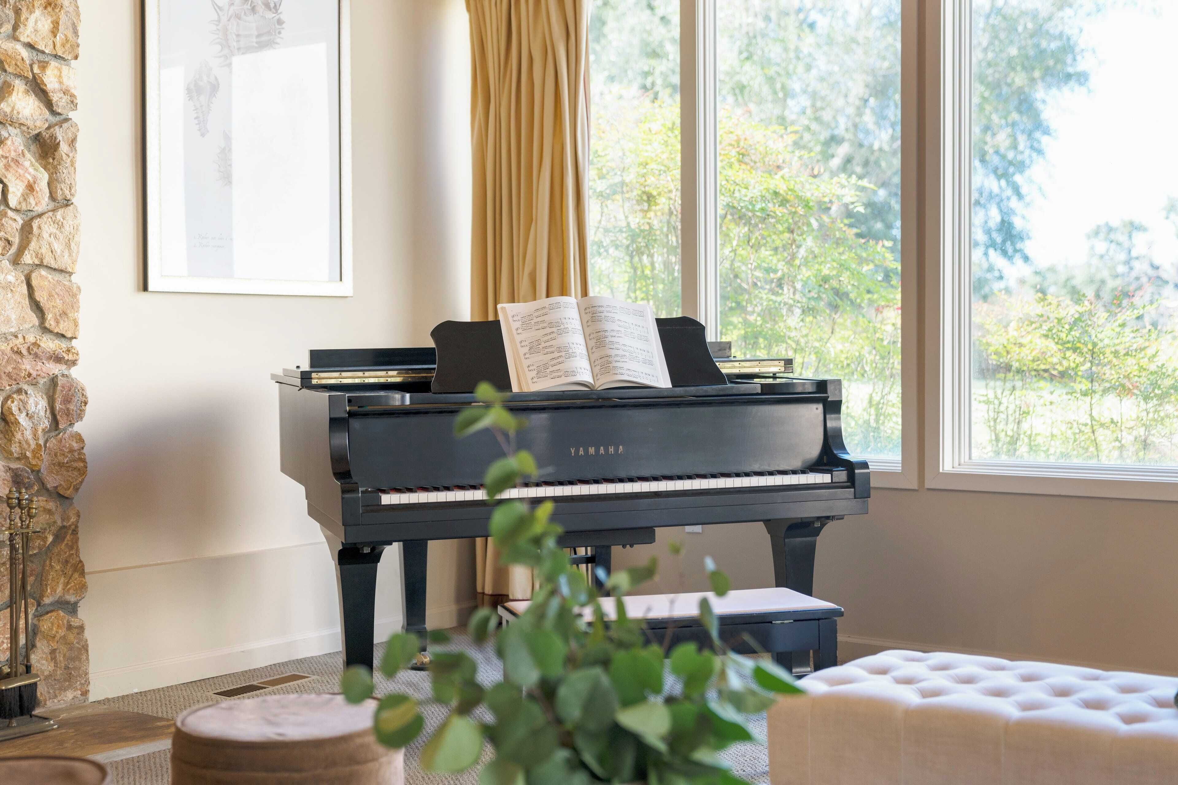 Een vleugelpiano met bladmuziek, een fotolijst aan de muur, en uitzicht op groen door grote ramen.