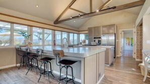 Una cocina moderna con una isla grande, taburetes de bar, electrodomésticos de acero inoxidable y amplias ventanas que ofrecen vistas.