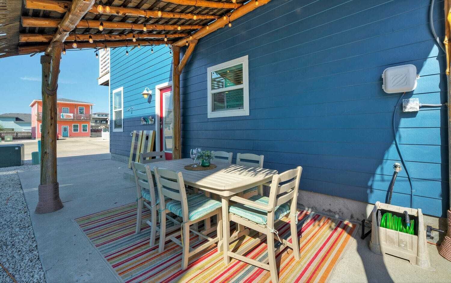 Een terras buiten met een houten tafel en stoelen, een kleurrijk gestreept kleed en een blauw gebouw op de achtergrond.