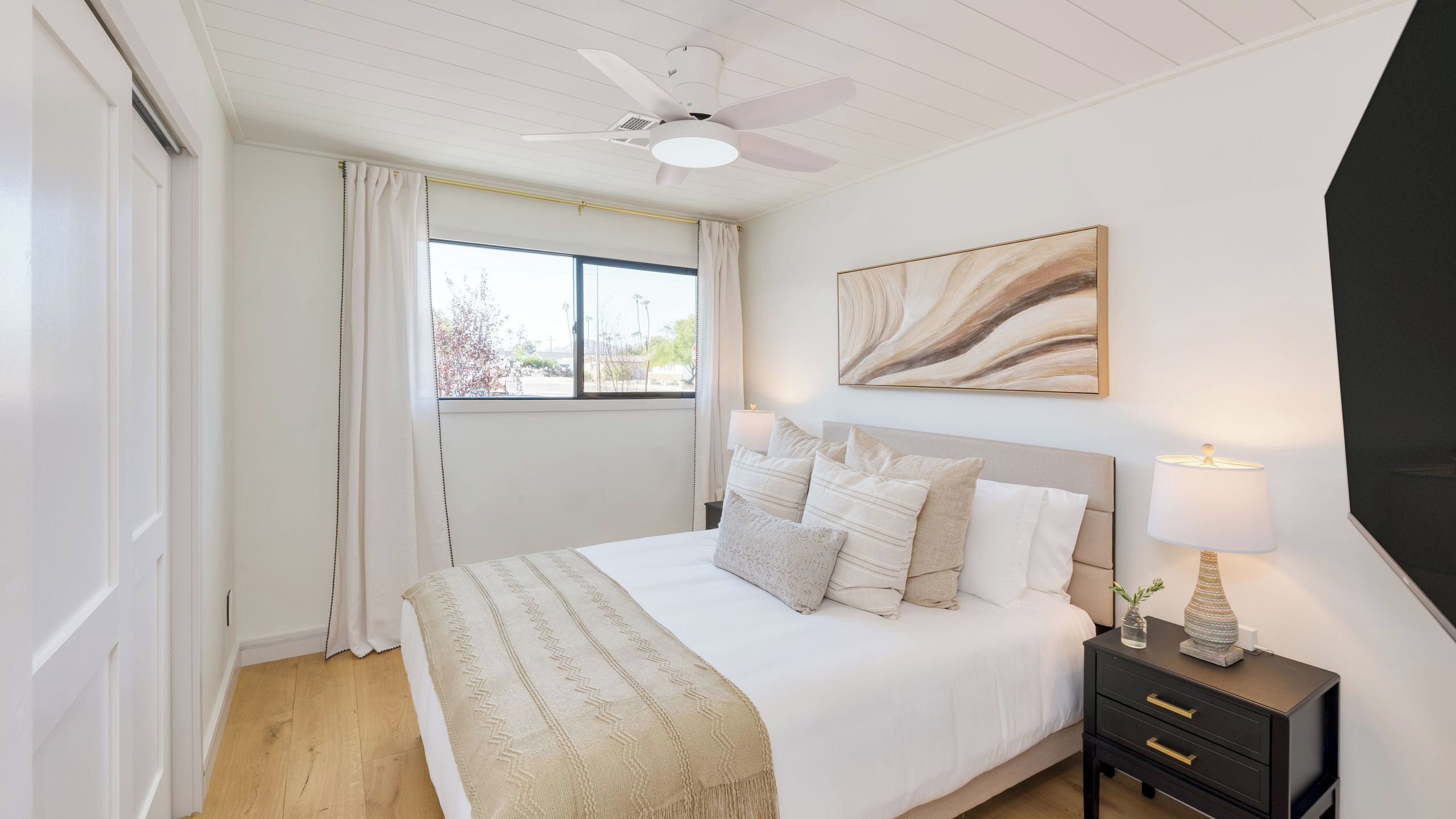 A bedroom with a bed, a nightstand, a ceiling fan, a window with curtains, and a painting on the wall.