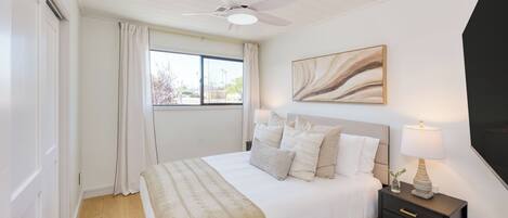 A bedroom with a bed, a nightstand, a ceiling fan, a window with curtains, and a painting on the wall.