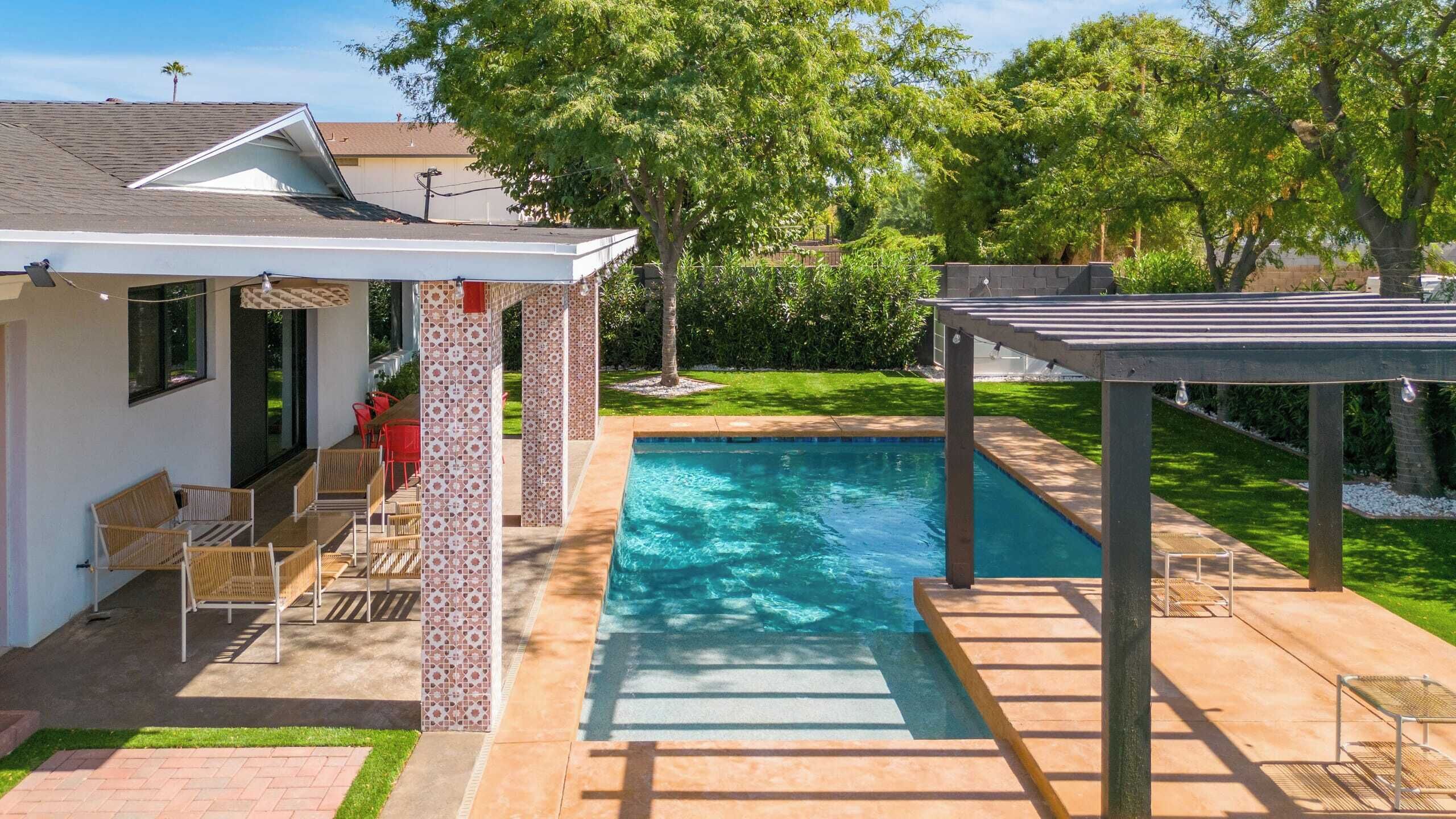 A swimming pool with a covered patio area, a small garden, and a house with a white exterior.