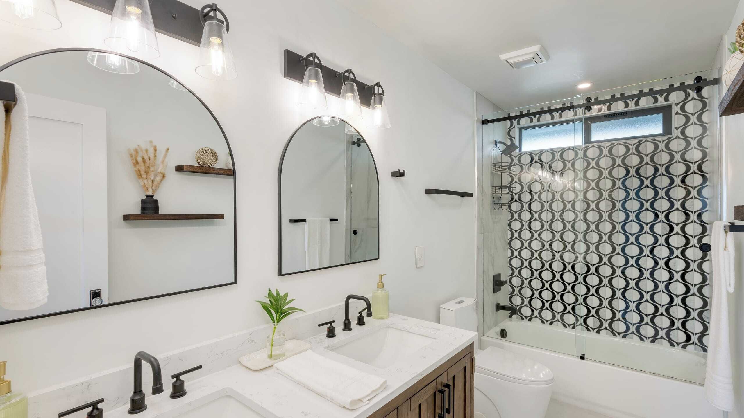 Een moderne badkamer met twee grote spiegels, een wastafel met een plant en een douche met een geometrisch tegelpatroon.