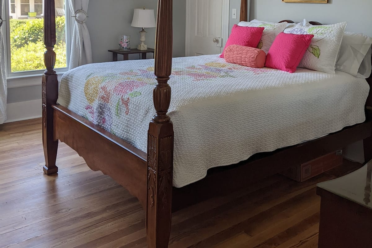 Ein Bett mit Baldachin, eine Blumendecke, ein Nachttisch mit Lampe und ein gerahmtes Blumenbild an der Wand.