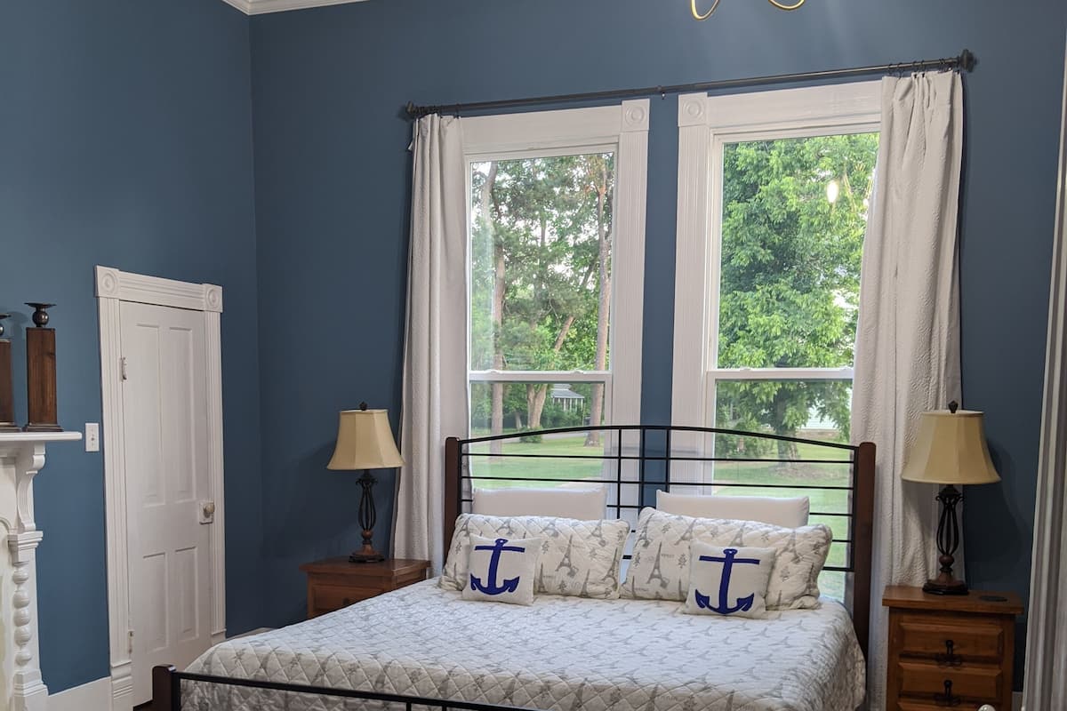 Ein Schlafzimmer mit einem Bett, zwei Nachttischen, einer Chandelüster, einem Fenster mit Vorhängen und Blick auf Grünfläche.