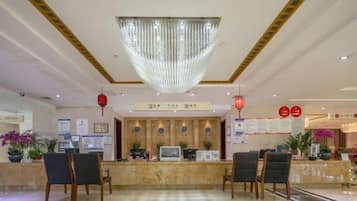 Uma área de recepção de hotel com lustre, balcão de atendimento, cadeiras e cartazes informativos.
