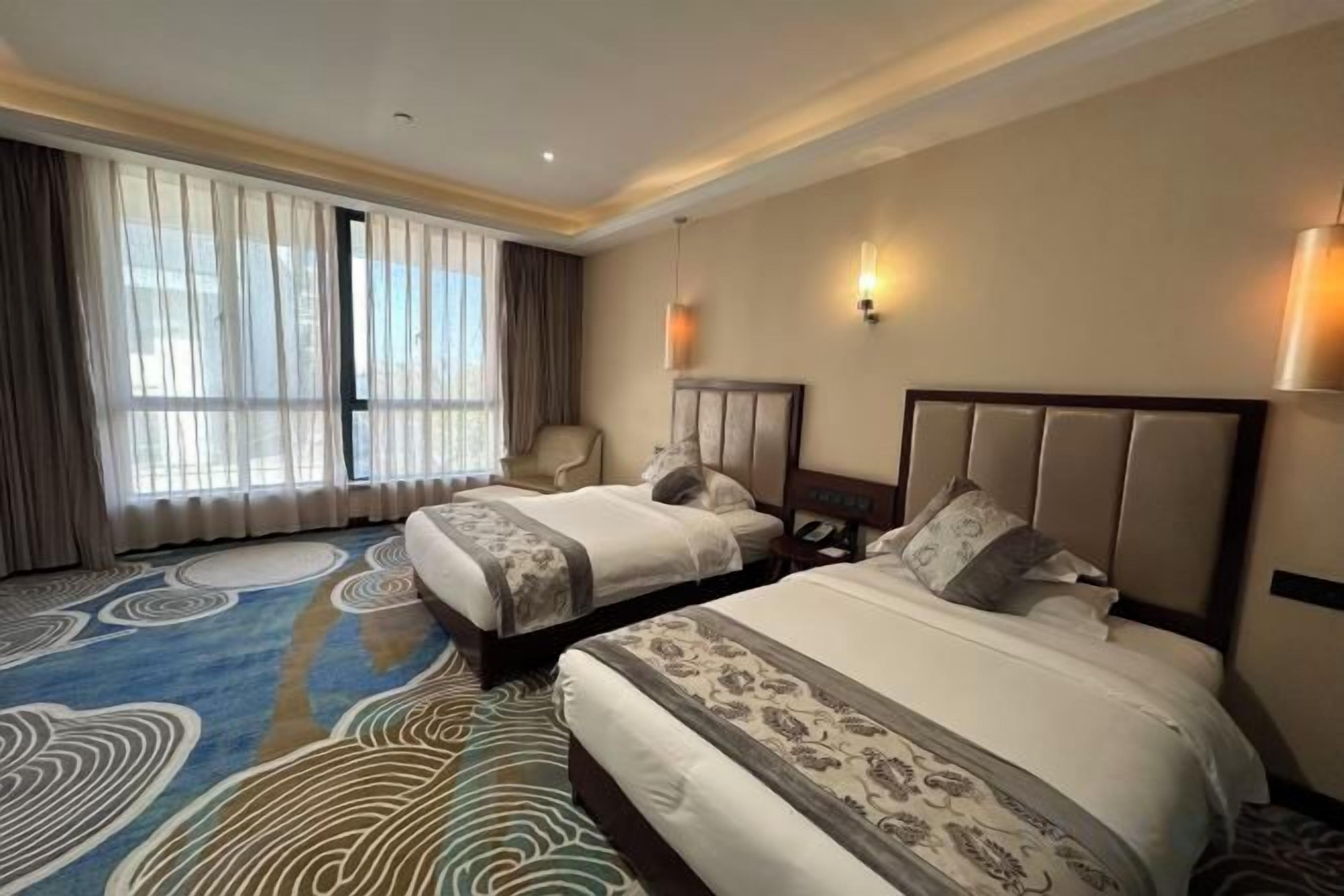 Een hotelkamer met twee bedden, een groot raam met gordijnen, een stoel en een tapijt met een rond patroon.