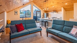 A modern loft with a wooden ceiling, large windows, a kitchen with white cabinets, and a blue sofa with cushions.