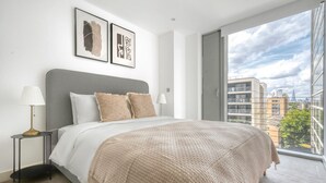 A modern bedroom with a large bed, two bedside tables, a view of the city through a large window, and two framed artworks on the wall. - Bright Canary Wharf 1BR, steps to South Quay light rail, by Blueground (London)