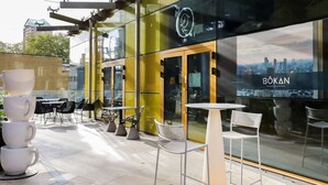 An outdoor seating area with tables and chairs, a glass wall with a cityscape view, and a sign for BOKAN restaurant. - Bright Canary Wharf 1BR, steps to South Quay light rail, by Blueground (London)