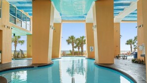 An indoor swimming pool with clear blue water, surrounded by beige walls and pillars, with a view of palm trees and a beach in the background.
