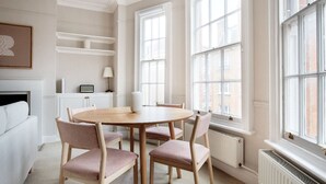 Une pièce avec une table ronde en bois et quatre chaises, de grandes fenêtres, un radiateur et une cheminée blanche.
