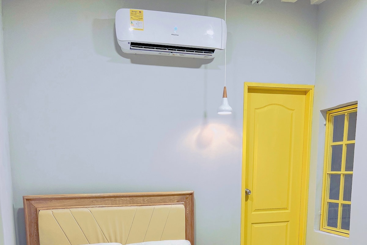 A neatly made bed with a wooden headboard, a small bedside table with a plant, and a yellow door with a window.