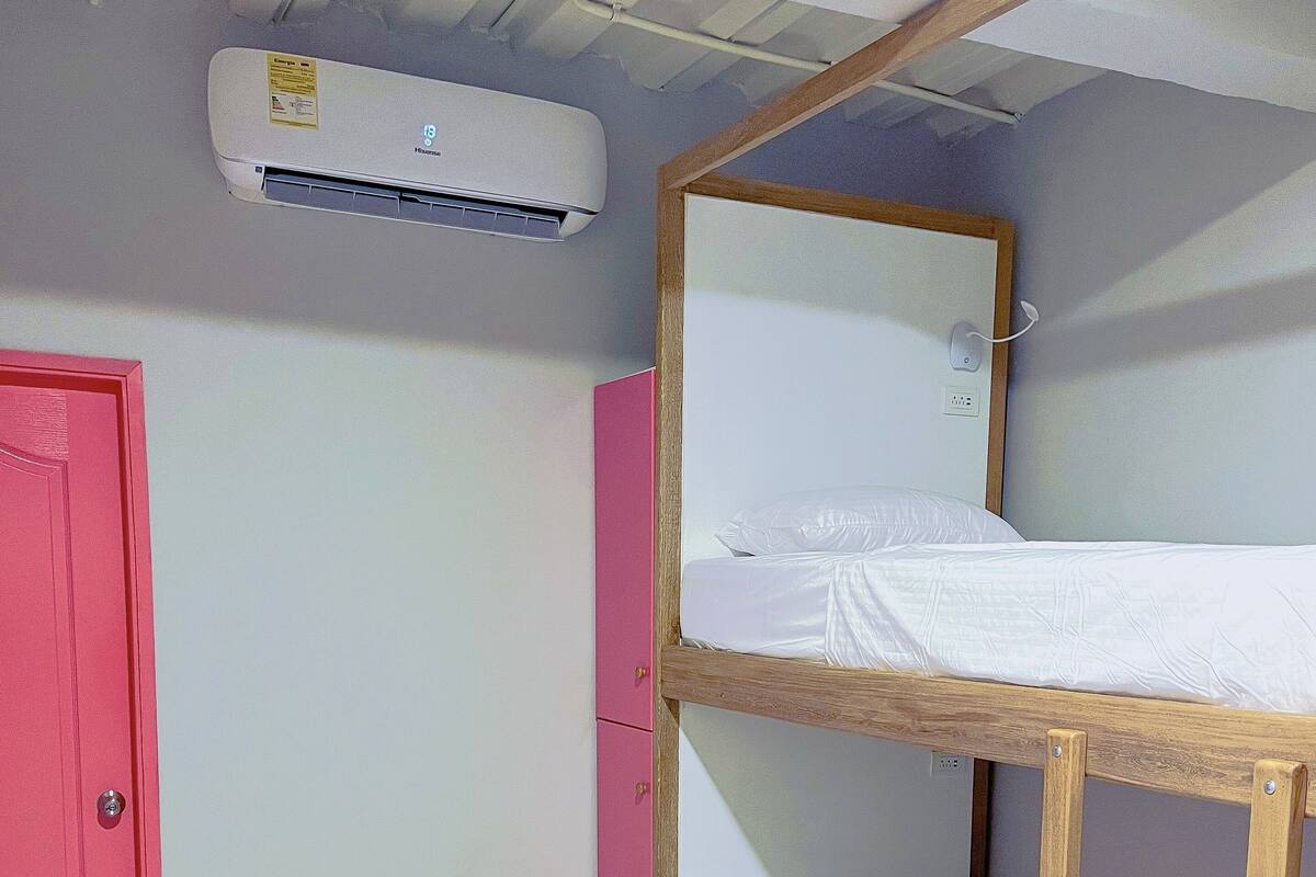 A bunk bed with a wooden frame and white bedding, a pink wardrobe, and an air conditioning unit on the wall.