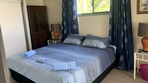 Ein ordentlich bezogenes Bett mit blauer Bettdecke und Kissen, ein Holzschrank, ein kleiner Tisch mit Lampe und ein Fenster mit Vorhängen.