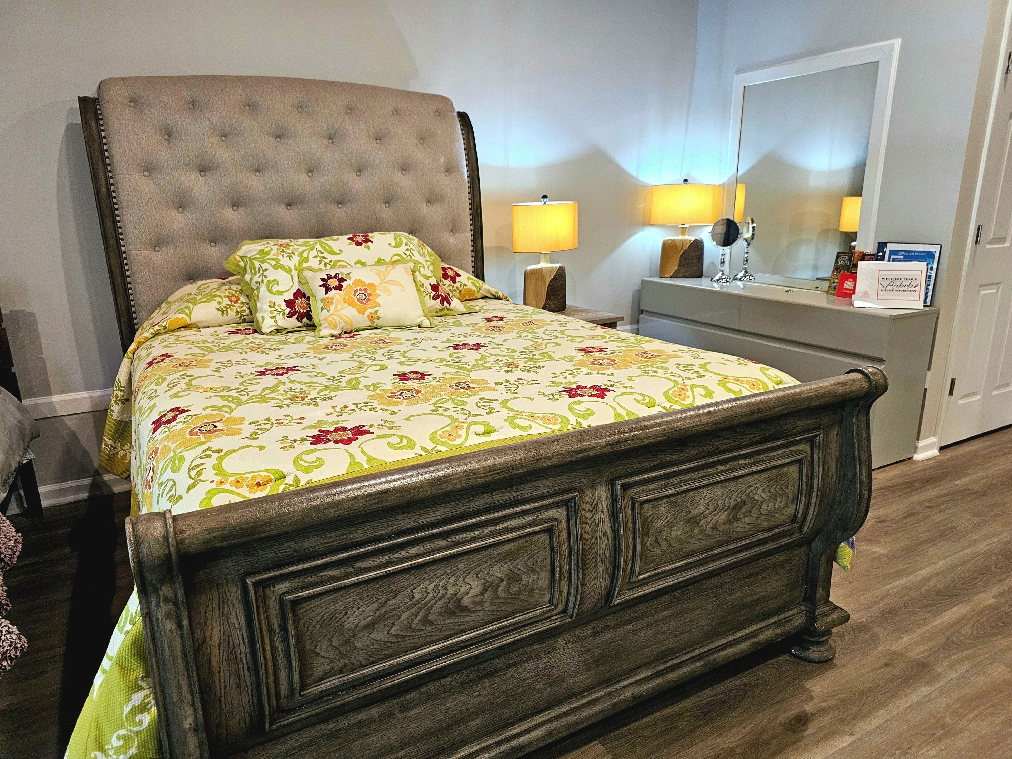 Un lit recouvert d’une couvre-lit à motifs floraux, une table de chevet avec des lampes et une commode avec un miroir.