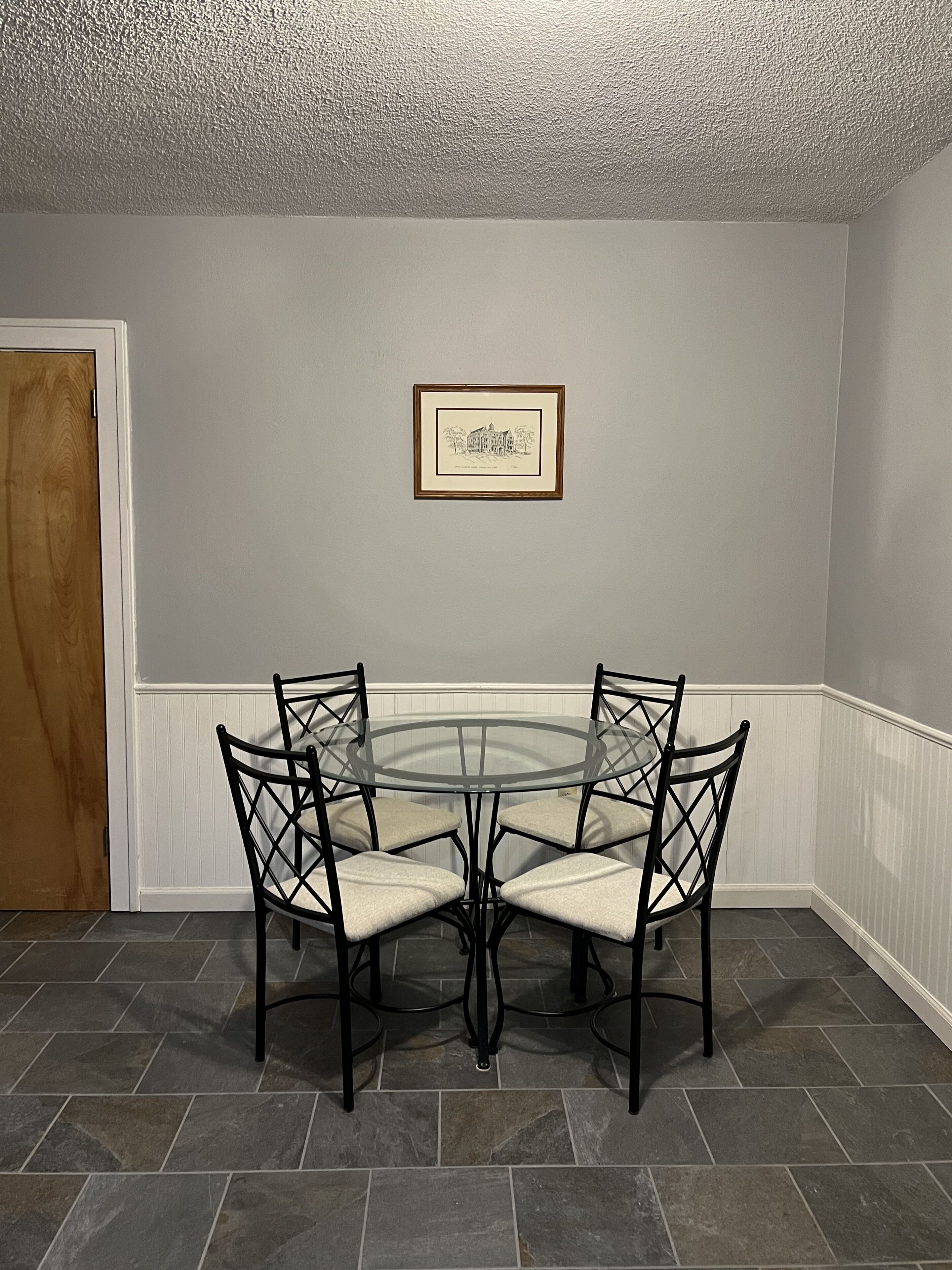 Un comedor con una mesa de vidrio y cuatro sillas, un cuadro en la pared, y una puerta a la izquierda.