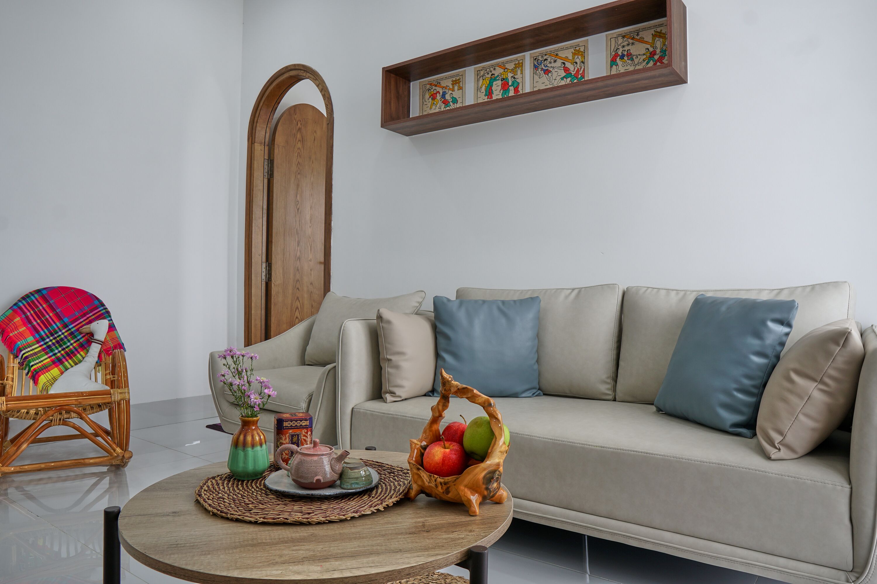 A living room with a sofa, a wicker chair, a round coffee table with a vase of flowers, and a wooden cabinet with artwork.
