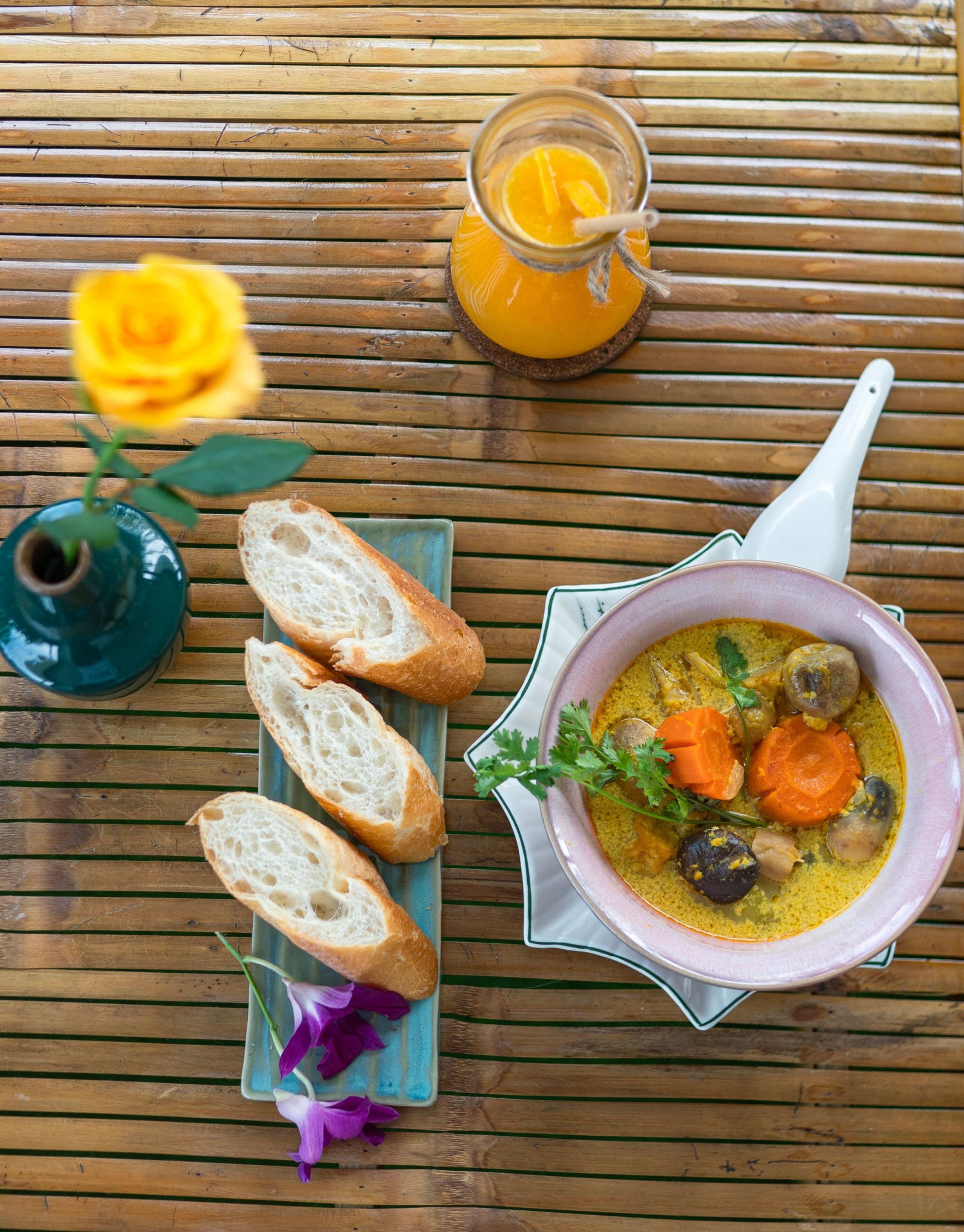 Ein Holztisch mit einer Schüssel Curry, Brotscheiben, einem Glas Orangensaft, einer Vase mit einer gelben Rose und einem Keramiklöffel.