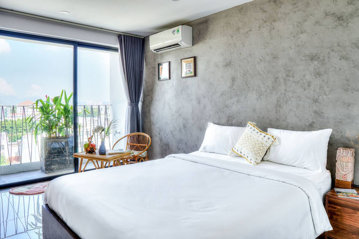 A modern hotel room with a large bed, a small table with chairs, a balcony with plants, and a concrete wall.
