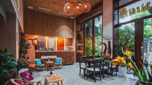 Un café acogedor con mobiliario de mimbre, un mostrador de recepción de madera y grandes ventanales con vistas a la calle.