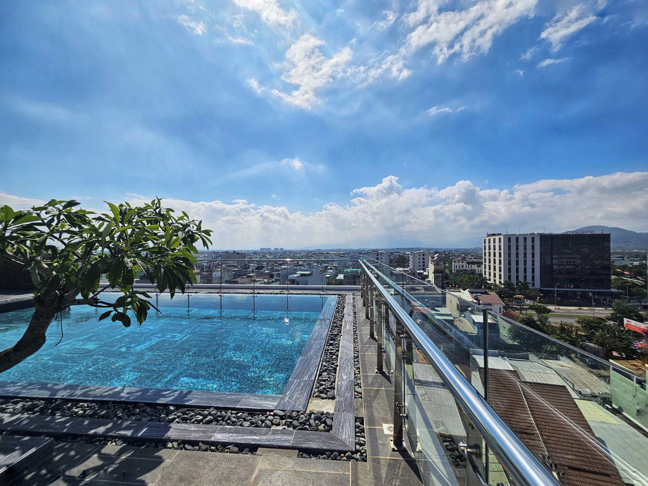 Una piscina en la azotea con barandilla de cristal, rodeada de baldosas de piedra y un árbol.