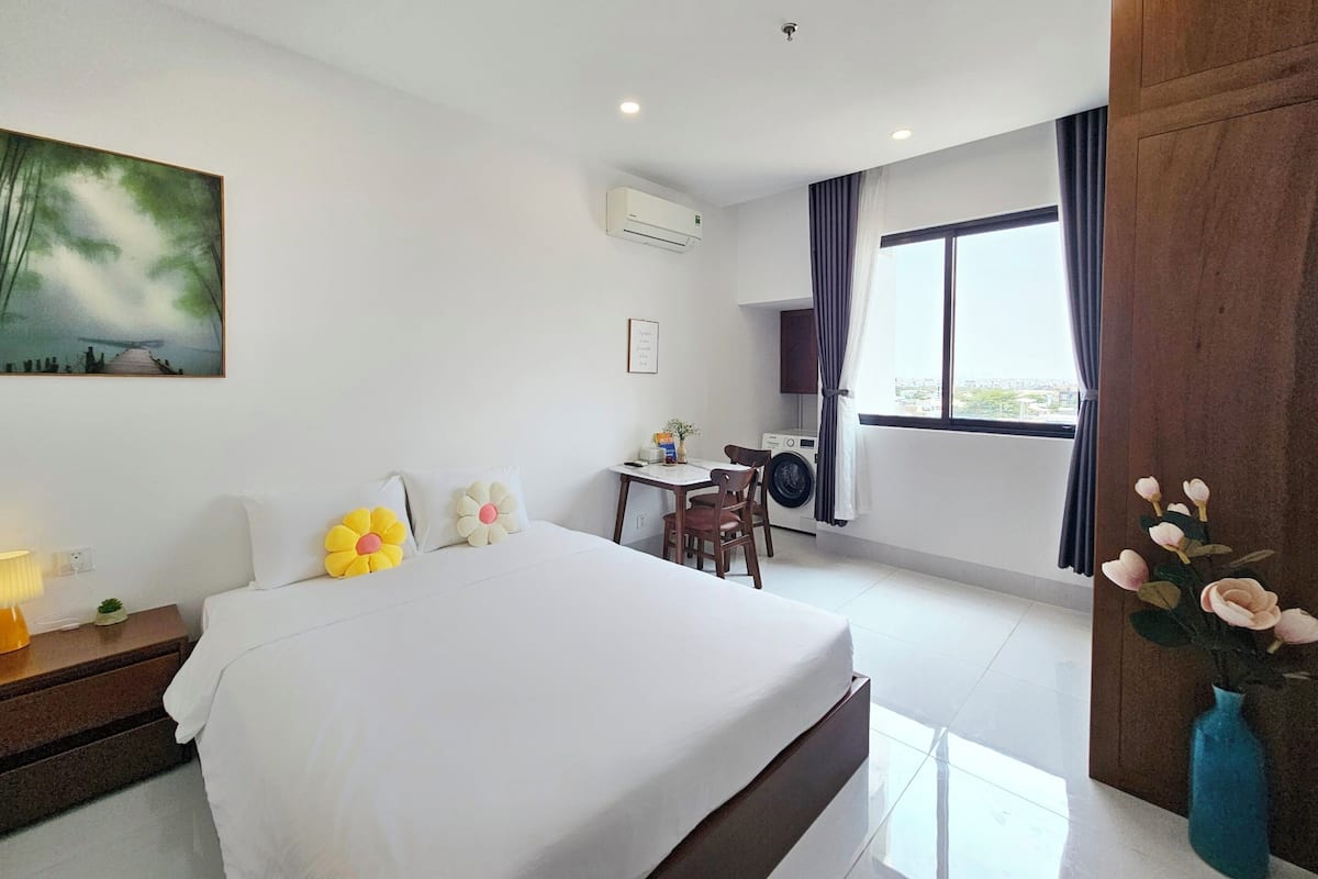 A hotel room with a bed, a desk with a chair, a washing machine, a window with curtains, and a painting on the wall.