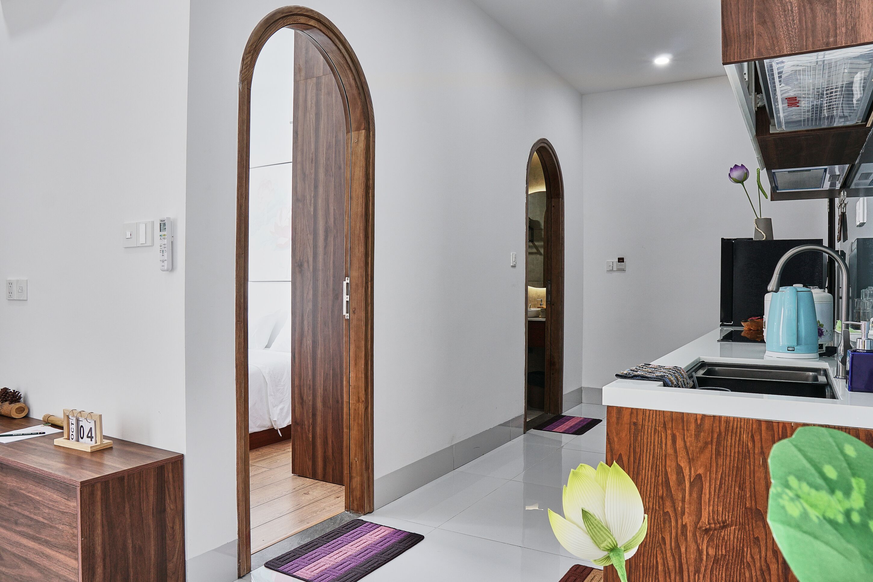 A modern hotel room with a wooden door, a small desk, a bed, a bathroom, and a kitchenette.