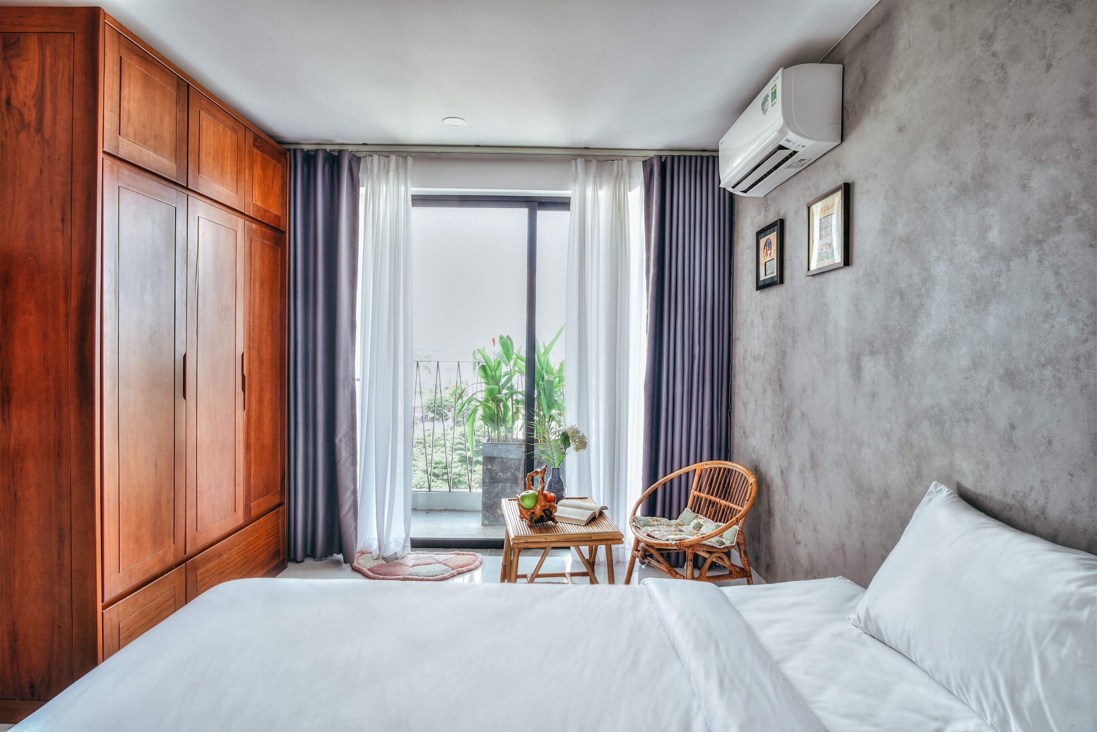 Une chambre d’hôtel avec un grand lit, une armoire en bois, une petite table avec un vase, un fauteuil en osier et une vue sur la verdure à l’extérieur.