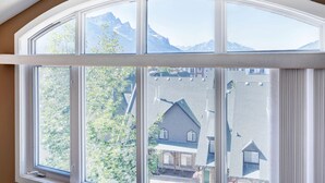 A large arched window with multiple panes, offering a view of a residential area with houses and mountains in the background. - Cozy Condo | Epic Mountain Views | Parking Garage | Central | Air Conditioning (Canmore)