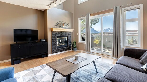A living room with a fireplace, a flat-screen TV, a sofa, and a large window with a view of mountains. - Cozy Condo | Epic Mountain Views | Parking Garage | Central | Air Conditioning (Canmore)