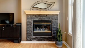 A living room with a fireplace, a television, and a potted plant. - Cozy Condo | Epic Mountain Views | Parking Garage | Central | Air Conditioning (Canmore)