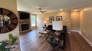 Un salon spacieux doté de planchers en bois franc, d’une cheminée en pierre, d’un ventilateur de plafond et d’un grand miroir.